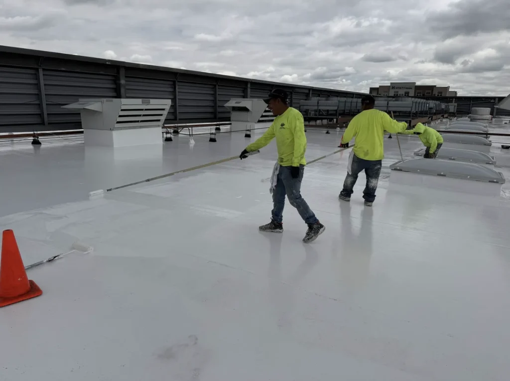 Two crew members in safety vests applying white coating to a commercial flat roof